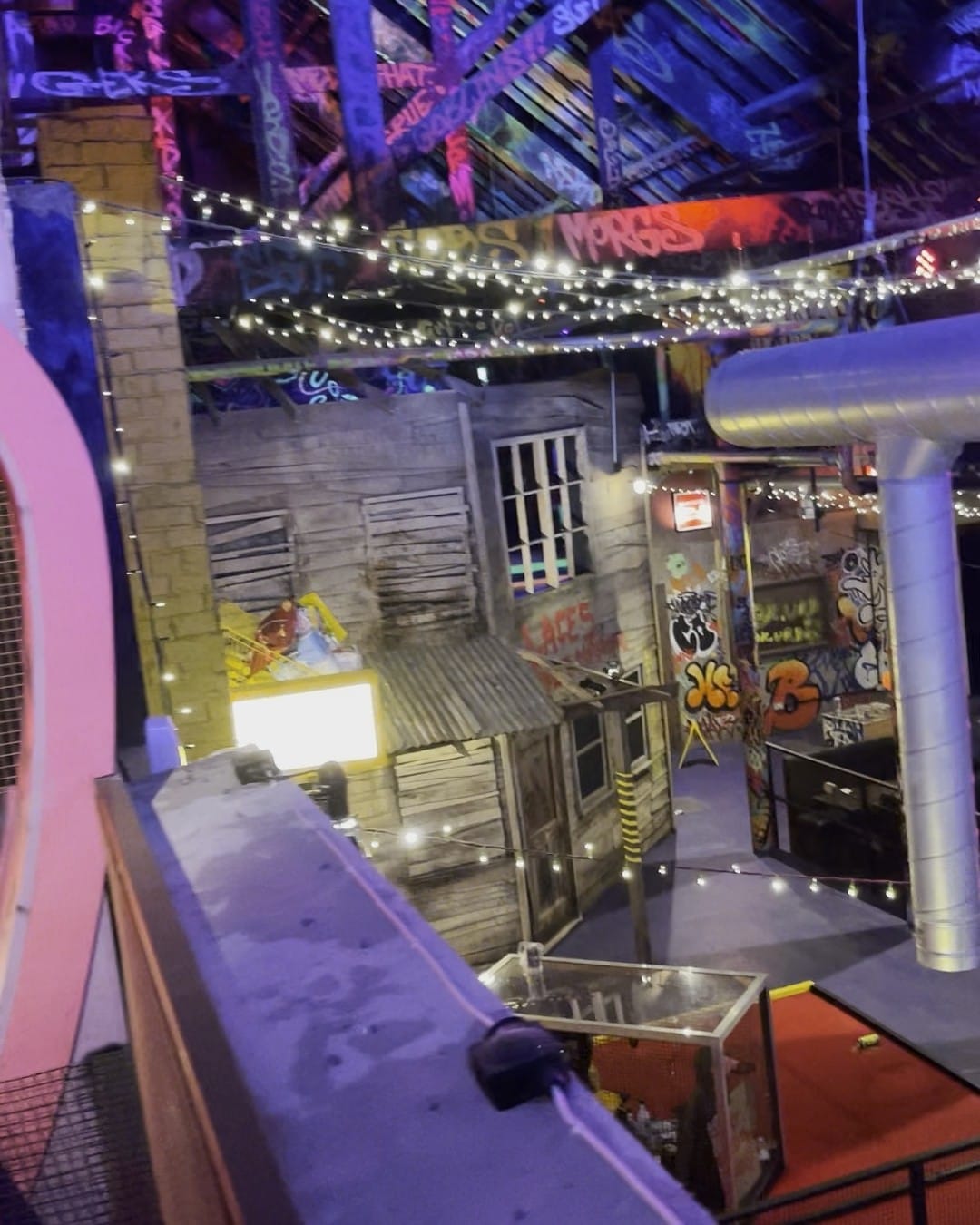 Looking down over the crazy golf course from a walkway: weathered timber shack fronts covered in graffiti, strings of fairy lights criss-crossing overhead, and a lit booth glowing at the far end.