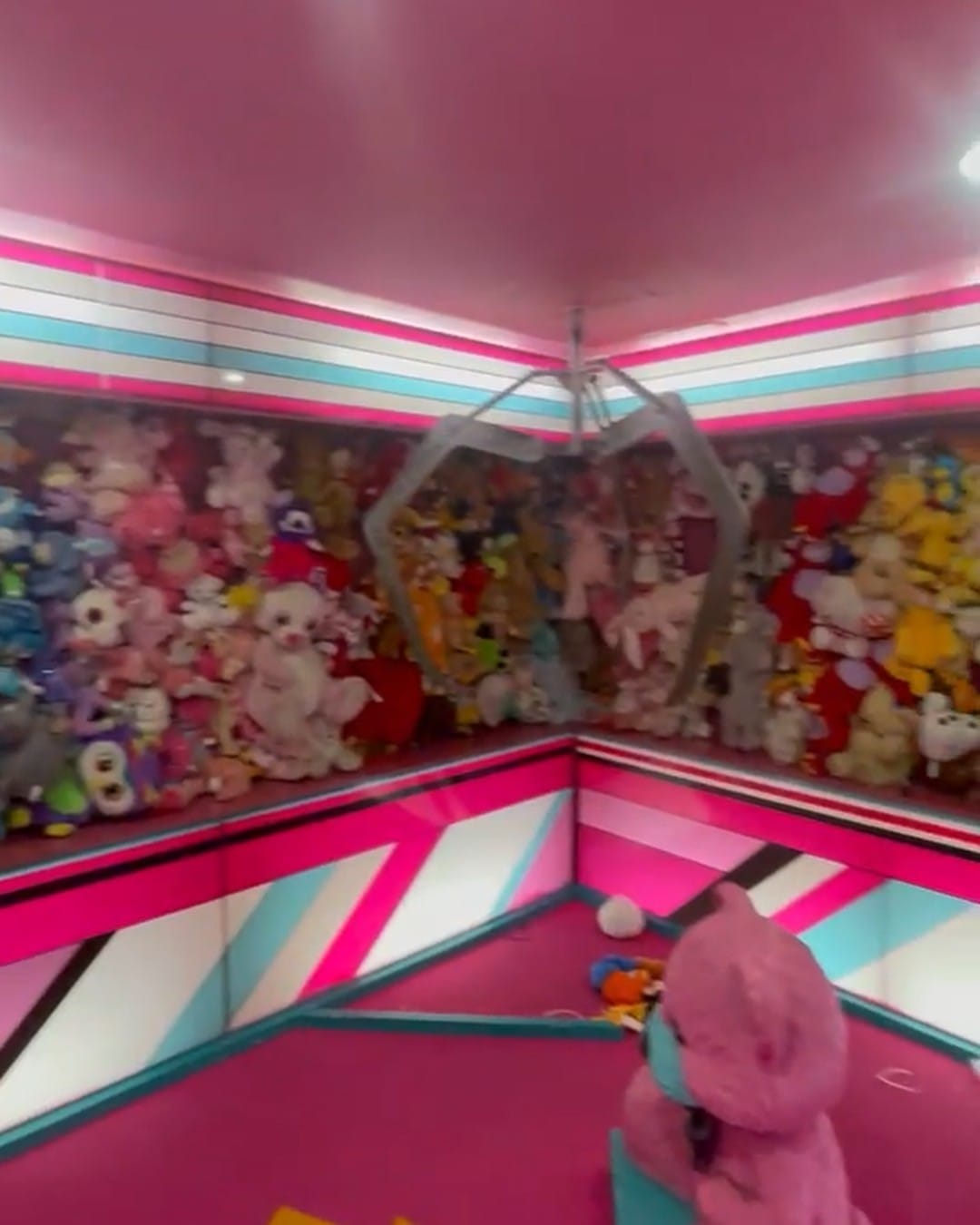 The inside of an oversized claw machine, pink and teal striped walls with a corner wall of soft toys and the grabber hanging in the middle.