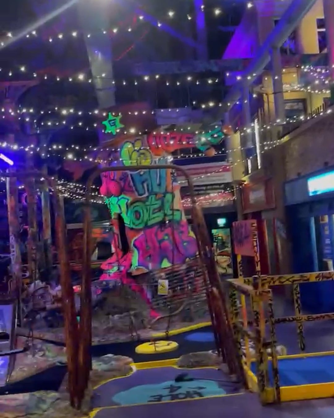 Hole four at ground level, a rusted metal archway painted in neon green and pink graffiti, mini golf greens underfoot and shopfronts lit up behind.