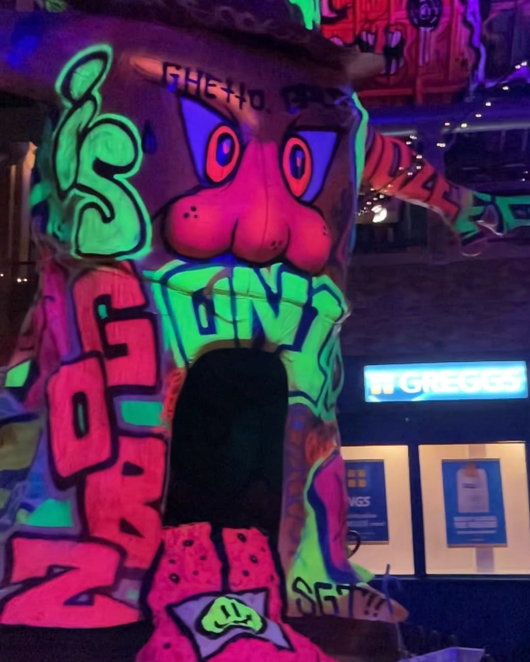 A huge neon mural of a cartoon face in green and pink painted across a pillar, with the lit sign of a mocked-up Greggs shopfront beside it.